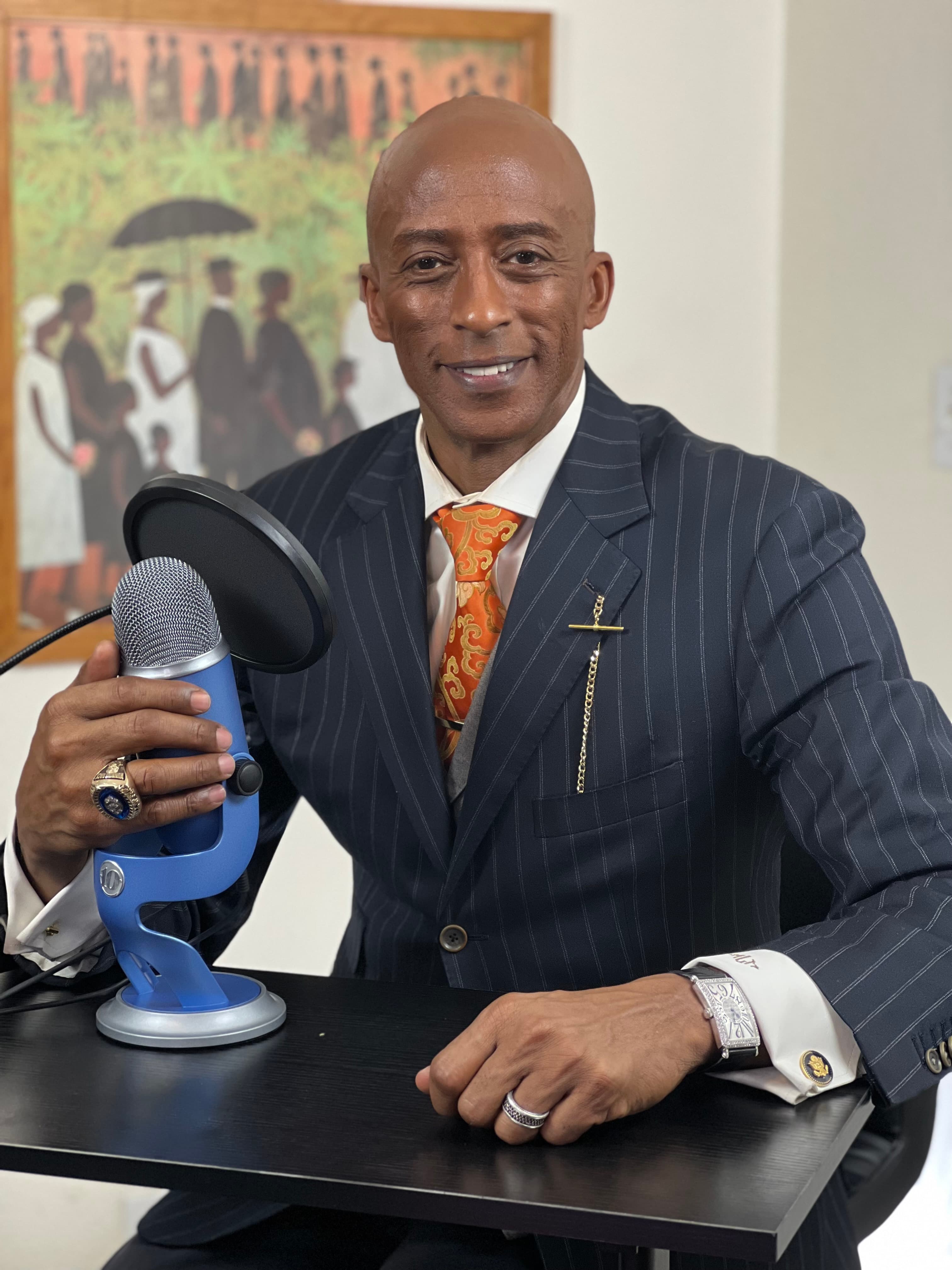 Smiling Black man in a pinstripe suit sits at a desk with a blue microphone.
